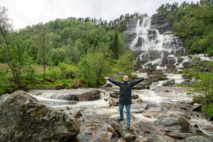 Tvindefossen Waterfall Voss Happy tourist with arms raised in front of the cascading Tvindefossen waterfall near Voss on a private tour with Jandis Fjord Tours.